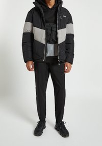 Giubbotto imbottito nero e grigio con cappuccio, dotato di zip frontale, pannelli a contrasto e tasca sul petto. Indossato sopra un hoodie grigio e pantaloni neri.