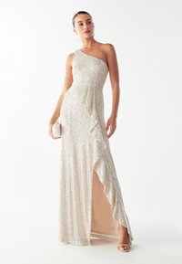 Vestido de noche con lentejuelas, diseño de un solo hombro, color beige, largo hasta el suelo con volantes laterales y una abertura alta, combinado con un pequeño bolso de mano.