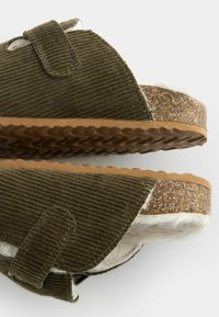 Sandales à glisser en velours côtelé vert olive avec des sangles ajustables, semelle en caoutchouc marron texturé et doublure blanche douce. Détails de couture notables visibles.