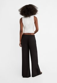 Kobieta z fryzurą afro, ubrana w biały top bez rękawów i luźne czarne spodnie, stojąca przodem do białego tła.