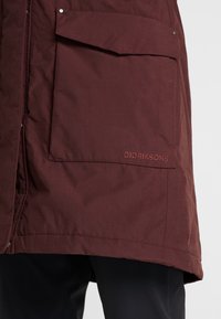 Burgundy vattentät parka med en texturerad tyg, stor framficka och diskret logotypdetalj på fållen. Skräddarsydd för en avslappnad passform.
