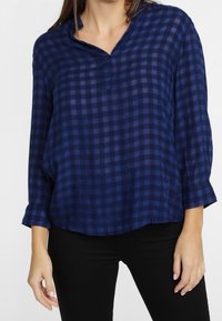 Kvinna iklädd en löst sittande långärmad skjorta i mörkblått och marinblått rutmönster tillsammans med svarta byxor mot en enfärgad bakgrund.