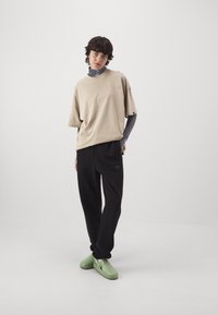 Beige oversized T-shirt met korte mouwen, zwarte casual broek met een relaxed fit, en lichtgroene klompen met uitsparingen. Geplaatst tegen een effen witte achtergrond.