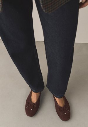 Person iført mørkeblå jeans og brune ballerinaer med sløjfer, stående på en lysegrå gulv.