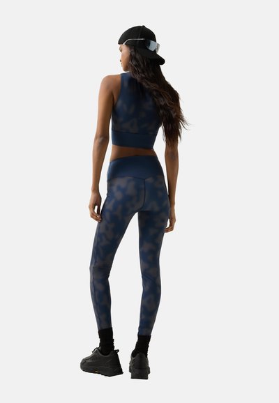 Kvinde iført marineblå camouflage leggings og crop top, sort kasket båret bagvendt, sorte sokker og sorte sneakers, stående med ryggen til mod en enkel baggrund.