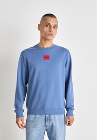 Felpa azzurra chiara con maniche lunghe e collo rotondo. Presenta una tasca logo rettangolare rossa sul petto. Tessuto morbido, vestibilità casual.