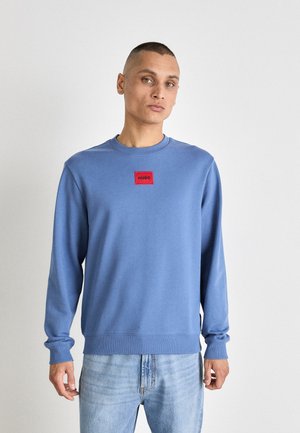 Lys blå genser med lange ermer og rund hals. Har et rødt rektangulært logo-patch på brystet. Mykt stoff, uformell passform.