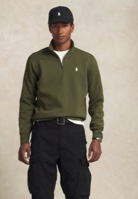 Sweatshirt verde-oliva com fecho, com um pequeno logo, usado sobre uma camisa branca, combinado com calças cargo pretas. Modelo a usar um chapéu preto.