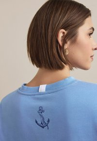 Modra bombažna jopa s okroglim izrezom, zanke na zadnjem vratu in vezenim mornarskim sidrom na hrbtu. Gladek tekstura.