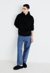 Sudadera de felpa negra con un bolsillo frontal, combinada con jeans rectos de un azul claro y zapatos slip-on de ante negros. Diseño minimalista.