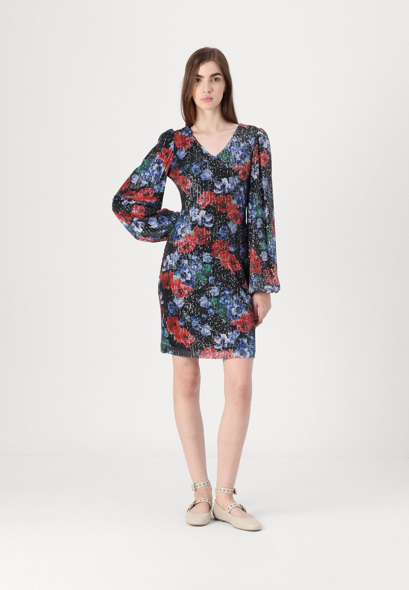 Robe noire à fleurs avec des motifs rouges, bleus et verts, dotée de manches bouffantes et d'un décolleté en V. Le modèle porte des chaussures à bride de cheville beiges.