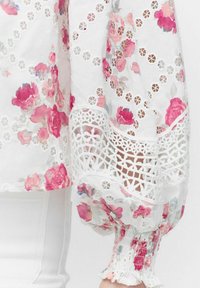Blomstret bluse med hvid bund, prydet med rosa og grå blomsterprint, blondekant og smockede manchetter. Blød, letvægtsstof, delikat design.