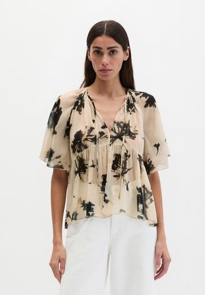 Frau trägt eine beige halbtransparente Bluse mit schwarzem Blumenmuster und lockeren kurzen Ärmeln, kombiniert mit weißen hoch taillierten Hosen.