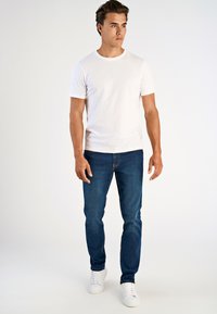 Valkoinen puuvillapaita, klassinen pyöreä pääntie, yhdistetty sinisiin farkkuihin ja valkoisiin tennareihin, seisoo neutraalilla taustalla.