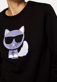 Gato morado con gafas de sol negras y collar con colgante, bordado en la parte frontal de un sudadera negra.