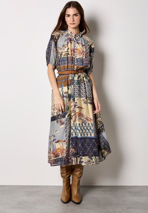 Jonge vrouw die een midi-jurk met patchwork-patroon draagt, korte mouwen, een riem en zandkleurige kniehoge laarzen, staand tegen een effen muur.