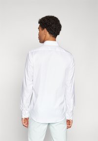 Camisa blanca de manga larga con botones y cuello clásico, que presenta una textura suave y un bajo curvado. Botones visibles en los puños.