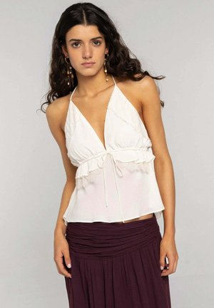 Mujer con cabello largo y oscuro que lleva un top halter blanco con encaje y una falda púrpura oscuro, de pie frente a un fondo claro y liso.