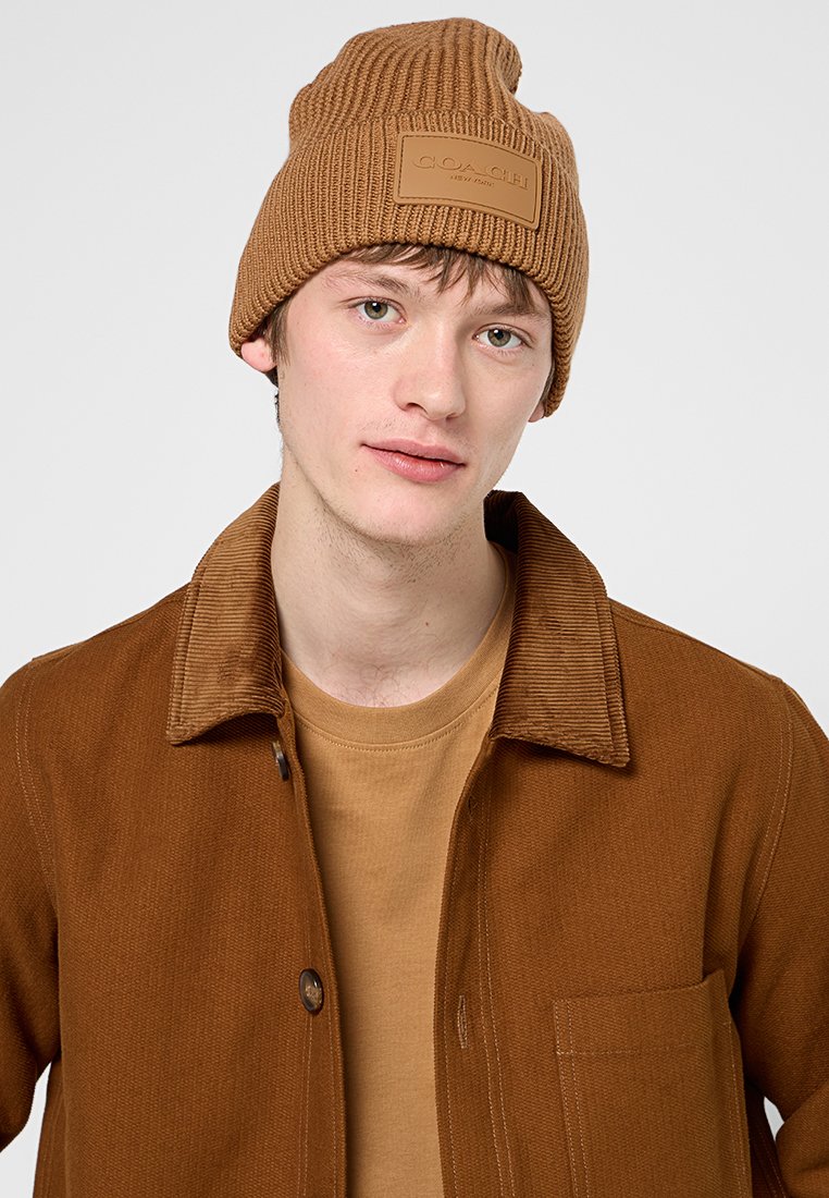 Ung man som bär en brun stickad mössa med en läderlogotyp, brun kordflodjacka och matchande brun tröja med rund hals.