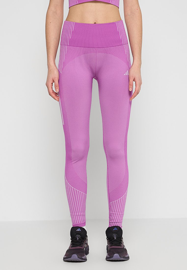 Legging de sport rose avec taille haute, finition texturée et rayures blanches verticales sur les côtés. Porté avec des chaussures de sport noires.