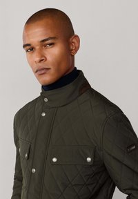 Jakna od maslinasto zelene tkanine s prešivanjem, koja ima visoki ovratnik, srebrne dugmadi s preklopom i dvije džepove na prsima. Uključuje smeđi naglasak na ovratniku.