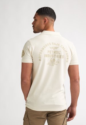 Polo color crema con maniche corte, caratterizzata da un grande logo stampato in oro sul retro e da un piccolo emblema sulla manica sinistra. Vestibilità casual.