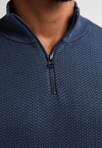 Pullover blu navy con zip, caratterizzato da un motivo zigzag testurizzato, con colletto alto e zip nera. Materiale morbido a maglia.