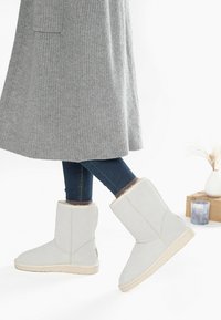 Botas de tobillo de color gris claro con una textura suave, forro de piel sintética y suelas de goma beige, combinadas con jeans oscuros y un abrigo largo de punto gris.