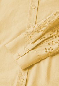 Camicia gialla con colletto abbottonato e dettagli ricamati intricati sulle maniche lunghe. Tessuto morbido con una texture liscia.