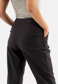 Pantalones de talle oscuro, ajustados, con una textura suave, que cuentan con bolsillos traseros y una cintura alta, estilizados con las manos en los bolsillos.