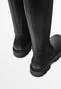 Bottes noires en cuir montant jusqu'au genou avec une finition lisse, présentant des coutures visibles et une semelle en caoutchouc épaisse et texturée.