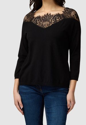 Haut noir à manches longues avec un col et un empiècement en dentelle florale transparente, associé à un jean en denim bleu, porté par une personne debout.