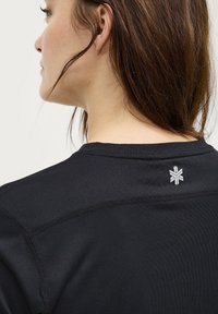 Schwarzes Sportshirt mit einer enganliegenden Passform, strukturiertem Stoff und einem kleinen weißen Logo nahe dem Kragen. Nahtdetails sind an den Schultern sichtbar.