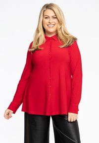 Chemise rouge à manches longues avec col, présentant une coupe ample et une texture lisse, associée à un pantalon en cuir noir.