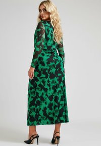 Pitkä vihreä mekko mustilla kukkakuviolla, läpinäkyvät pitkät hihat ja vartalonmyötäinen siluetti. Kangas näyttää kevyeltä ja virtaavalta.