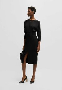 Svart midiklänning i mjukt material, med lång ärm, en slits fram och midja med bälte. Bärs med svarta klackpumps och en handväska.