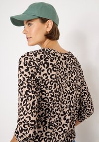 Top in stampa leopardata beige con macchie nere, con maniche a tre quarti. La modella indossa un cappellino da baseball verde. Texture morbida.