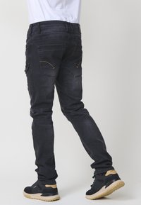 Vaqueros de mezclilla negros con un corte ajustado, que presentan detalles en costura dorada y bolsillos tipo parche, combinados con zapatillas negras sobre un fondo claro.