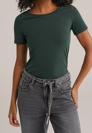 Vrouw draagt een nauwsluitende donkergroene korte mouw top, ingestopt in een hooggetailleerde grijze spijkerbroek met een bijpassende stoffen riem die vooraan is gestrikt.