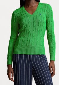 Suéter verde de punto trenzado con diseño de escote en V, puños y dobladillo acanalados. Presenta un pequeño acento de logo. Combinado con pantalones a rayas azul marino.