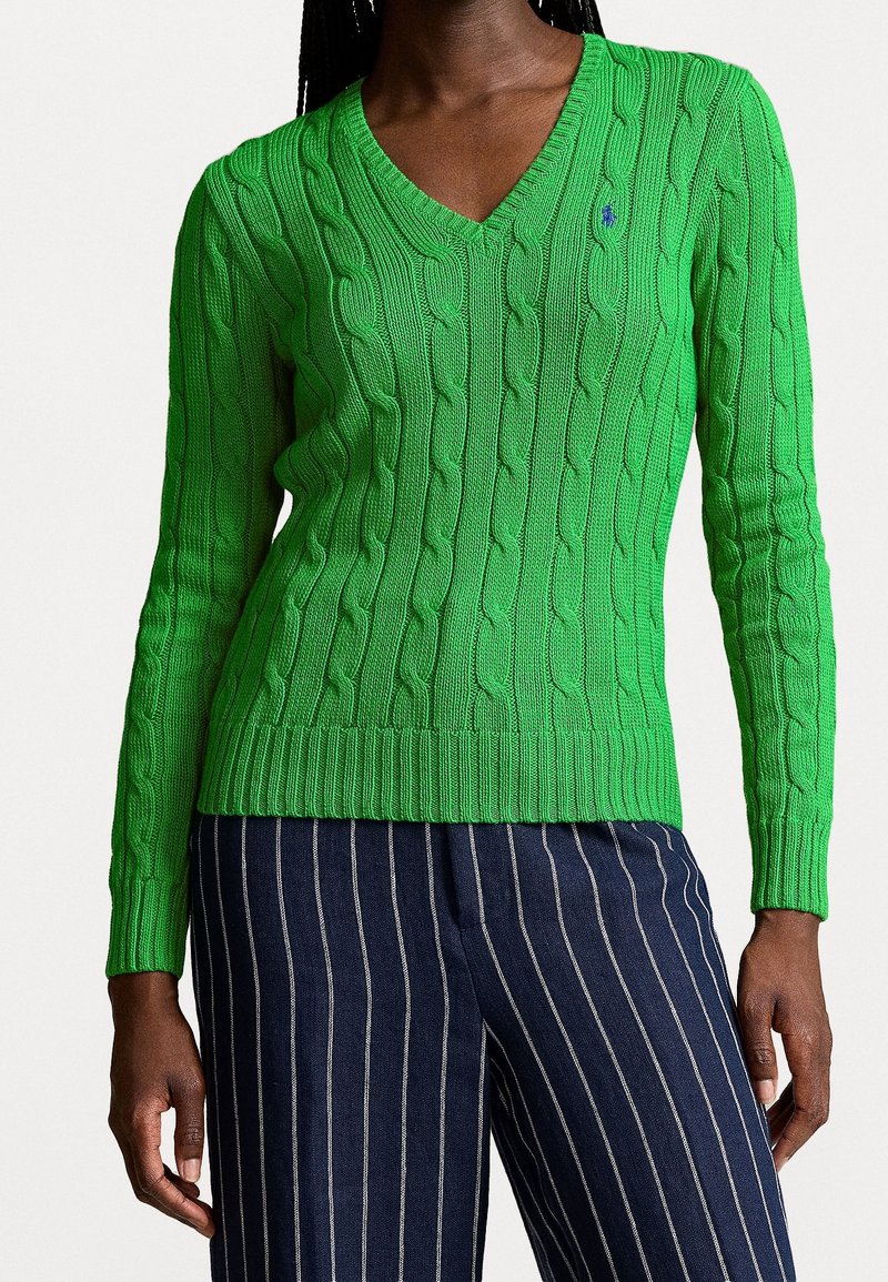 Suéter verde de punto trenzado con diseño de escote en V, puños y dobladillo acanalados. Presenta un pequeño acento de logo. Combinado con pantalones a rayas azul marino.