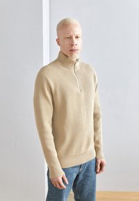 Maglione beige a coste con colletto alto e zip a metà, abbinato a jeans in denim blu. Il tessuto appare texture e morbido.