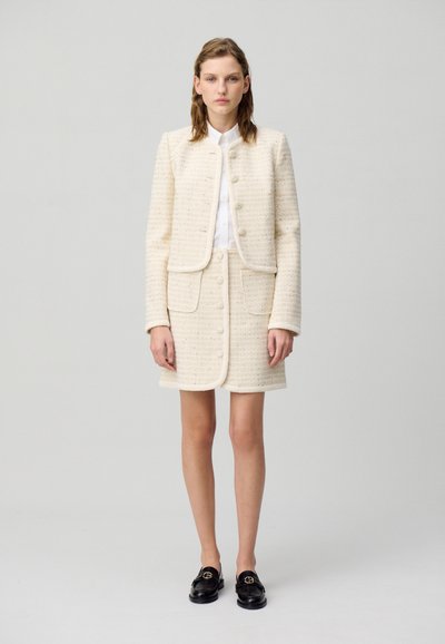 Un costume en tweed blanc avec une veste ajustée et une jupe courte, présentant de grands boutons et une finition texturée. Associé à des mocassins noirs.