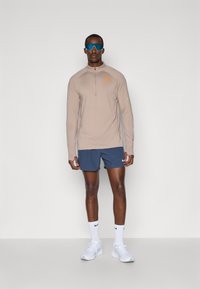 Cămașă sport cu mânecă lungă, de un marron deschis, cu fermoar și logo portocaliu, purtată cu pantaloni scurți albaștri și adidași albi. Ochelari de soare sport incluși.