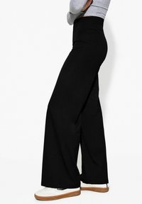 Pantalons larges noirs en tissu lisse, dotés d'une taille haute et d'une finition nette au bas, associés à des baskets blanches.
