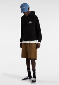Zwarte hoodie met logo, witte lange mouwen shirt eronder, bruine shorts, zwarte sneakers en blauwe pet met geborduurd logo detail.