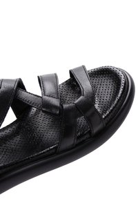 Black leather sandal with crisscrossed straps, perforated insole, and a rounded toe. Smooth texture with a flexible rubber sole.