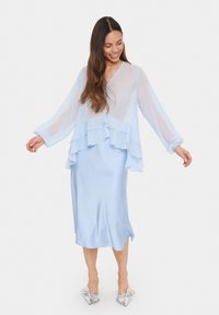 Abito a strati di colore azzurro chiaro con maniche lunghe in tessuto trasparente e finitura simile alla seta. Scarpe a ballerina con fiocco argentato completano l'outfit.