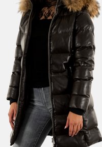 Abrigo acolchado de color marrón, con capucha de pelo sintético, acabado brillante, cierre con cremallera y puños negros; modelo que lleva vaqueros grises y una camiseta negra.