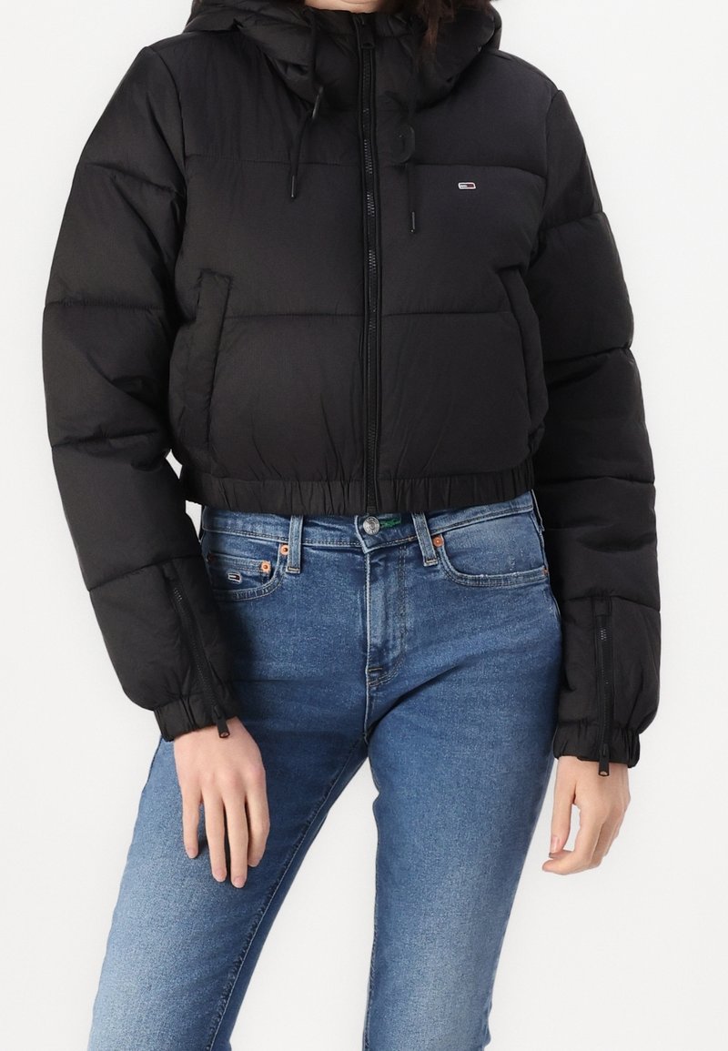 Czarna krótka puchowa kurtka z kapturem, z zamkiem błyskawicznym z przodu, elastycznym dołem i bocznymi kieszeniami. Nosi się na niebieskich dżinsach. Miękka faktura, izolacyjny design.